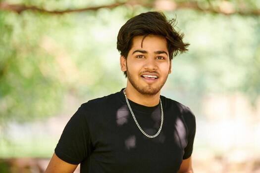 smiling-young-indian-man-portrait-in-black-t-shirt-and-silver-neck-chain-outdoor-green-public-park-photo
